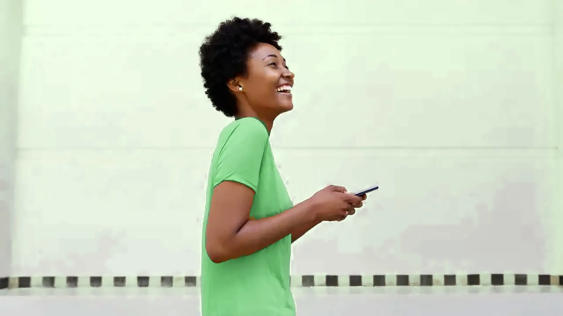 Jeune femme en t-shirt vert qui marche en riant, téléphone à la main.