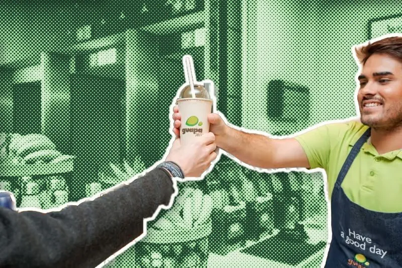 Un membre de l’équipe Guapa Juice tend un jus frais à un client par-dessus le comptoir, sur une trame de points verte.