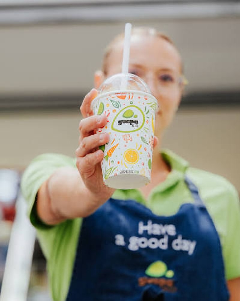 Une employée de Guapa Juice tend un jus au comptoir, tablier « Have a good day »