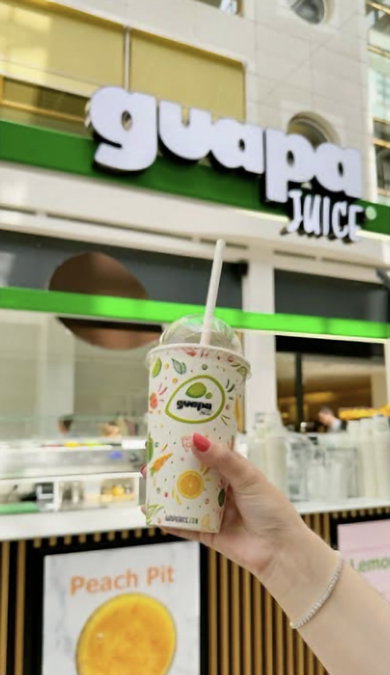 Un jus Guapa Juice tenu devant l'enseigne d'un bar à jus de la chaîne